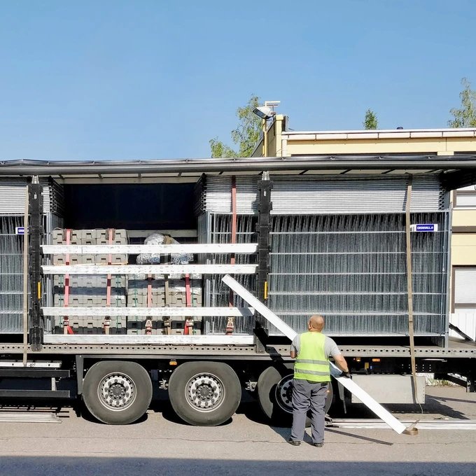 Ein Arbeiter mit Warnweste entlädt lange Metallstangen von einem LKW, der Metalizäune und Betonblöcke trasnportiert.