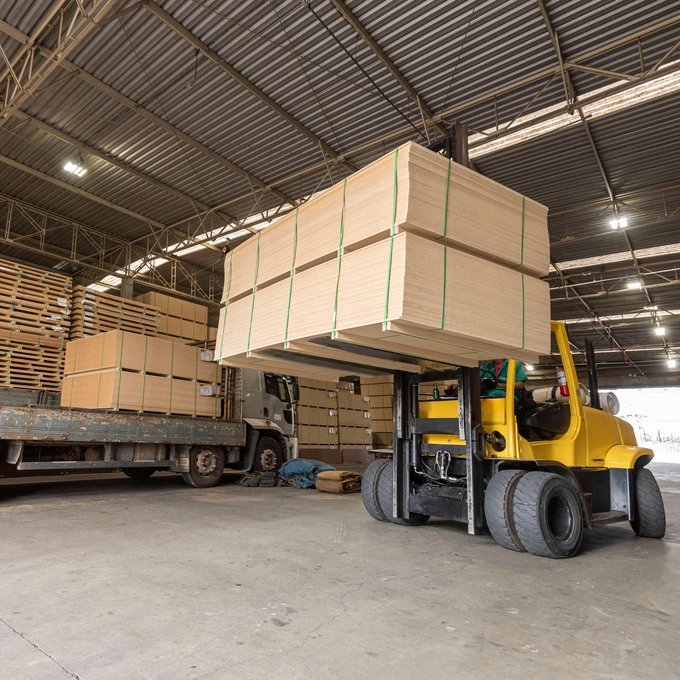 Ein gelber Gabelstapler hebt einen großen Stapel Sperrholzplatten in einem Lagerhaus, im Hintergrund sind weitere Sperrholzbündel und ein Lastwagen zu sehen.