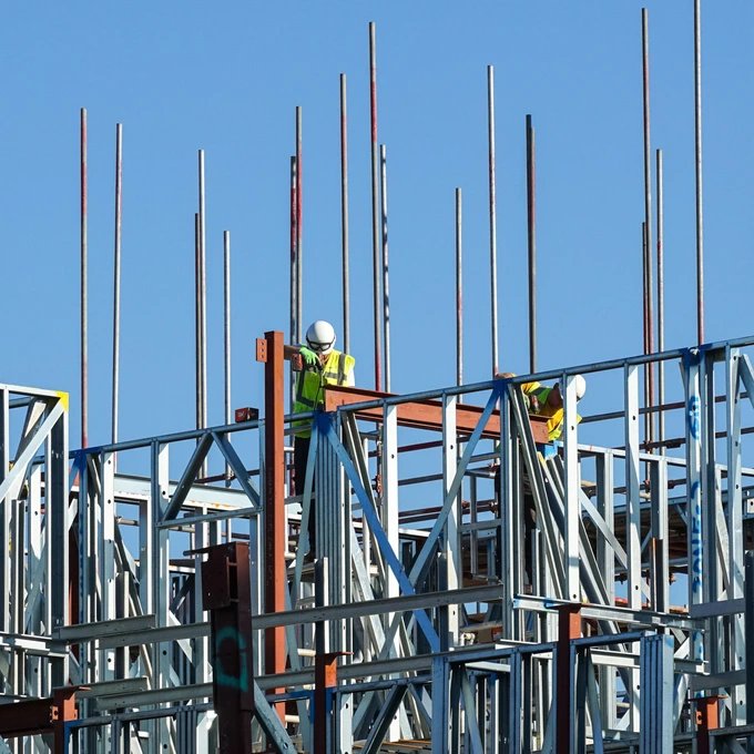 Bauarbeiter in Sicherheitskleidung arbeiten an einem Stahlrahmenbau auf einer Baustelle unter klarem blauen Himmel.